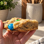 A hand holding a large, golden-brown cookie loaded with colorful candy-coated chocolate chips, set against a warm kitchen background with a potted plant and ceramic jar.