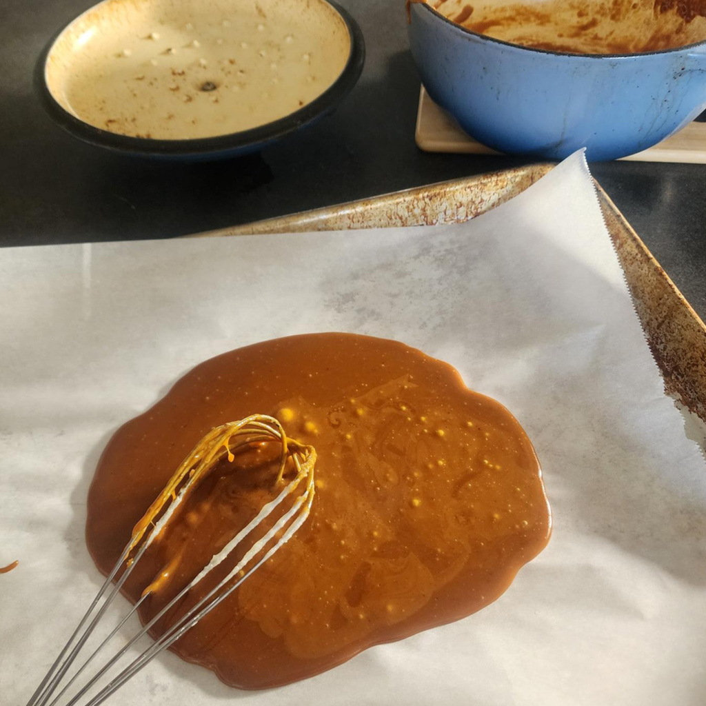 A close-up of caramel sauce being whisked on a baking sheet lined with parchment paper, with empty bowls in the background.