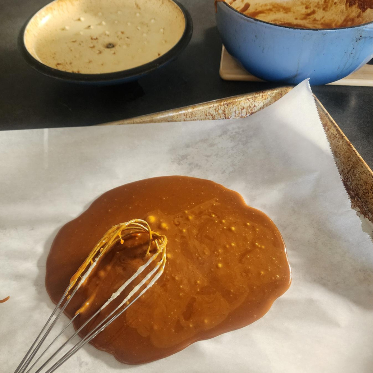 A close-up of caramel sauce being whisked on a baking sheet lined with parchment paper, with empty bowls in the background.